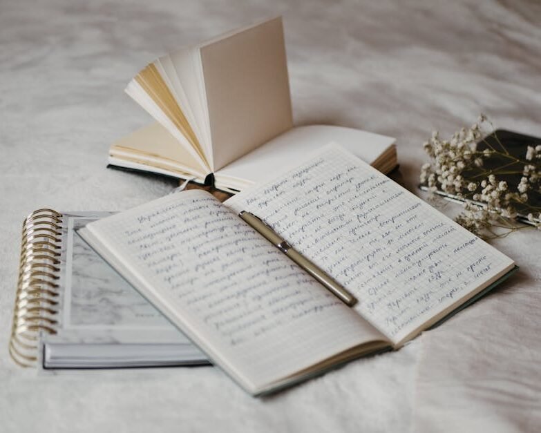 Close-up of open notebooks and a pen on a soft bed surface with delicate dried flowers.