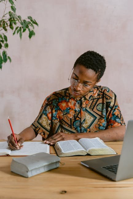 Person writing notes from a book with a laptop on a wooden table indoors.