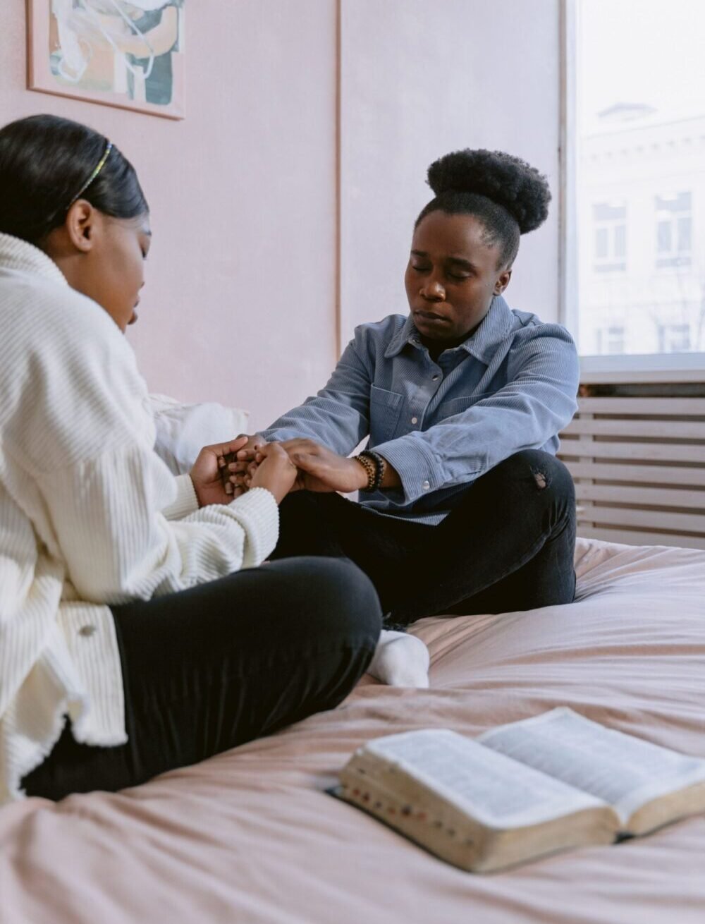 Two women in a bedroom, seated and praying together by an open Bible on a bed.